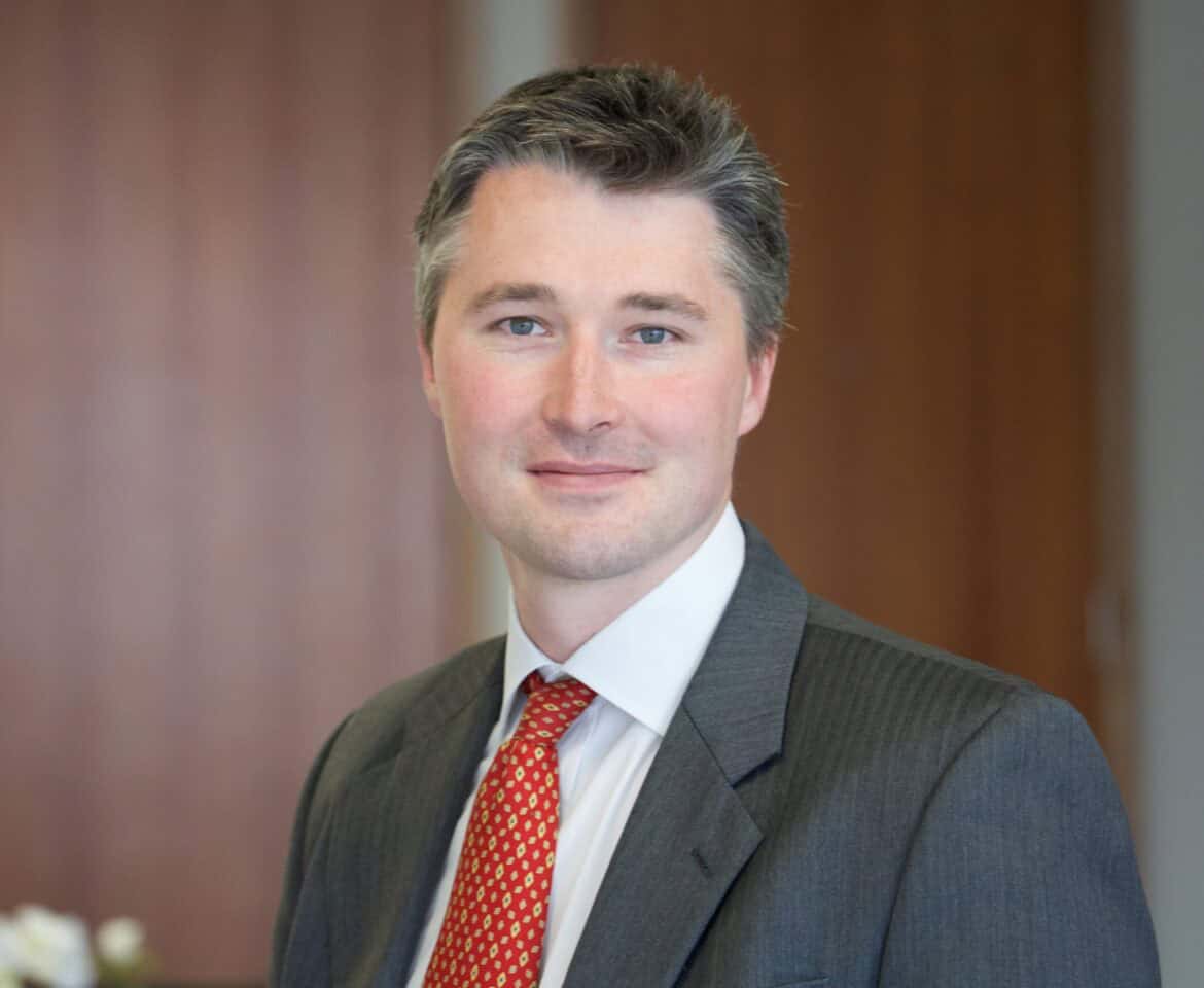 Headshot of a man with blue eyes and graying dark hair, wearing a gray suit and red patterned tie, smiling.