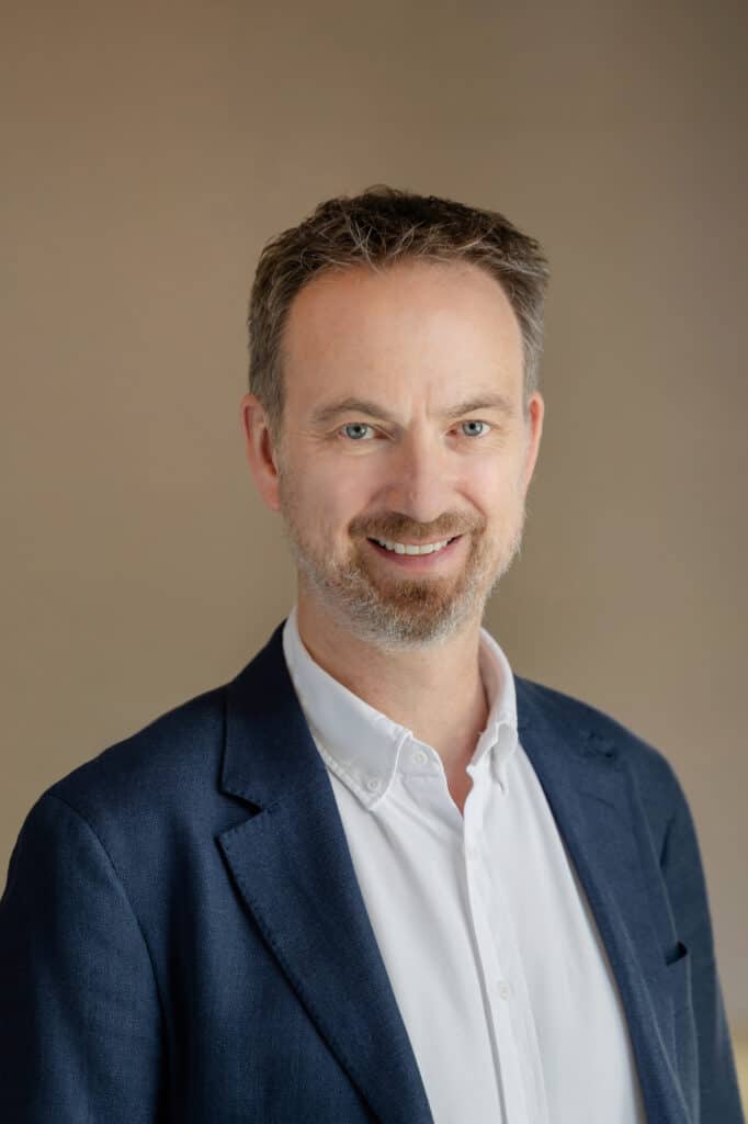 Headshot of a smiling man with blue eyes, short brown and grey hair, and a beard, wearing a white shirt and blue blazer against a light brown background.