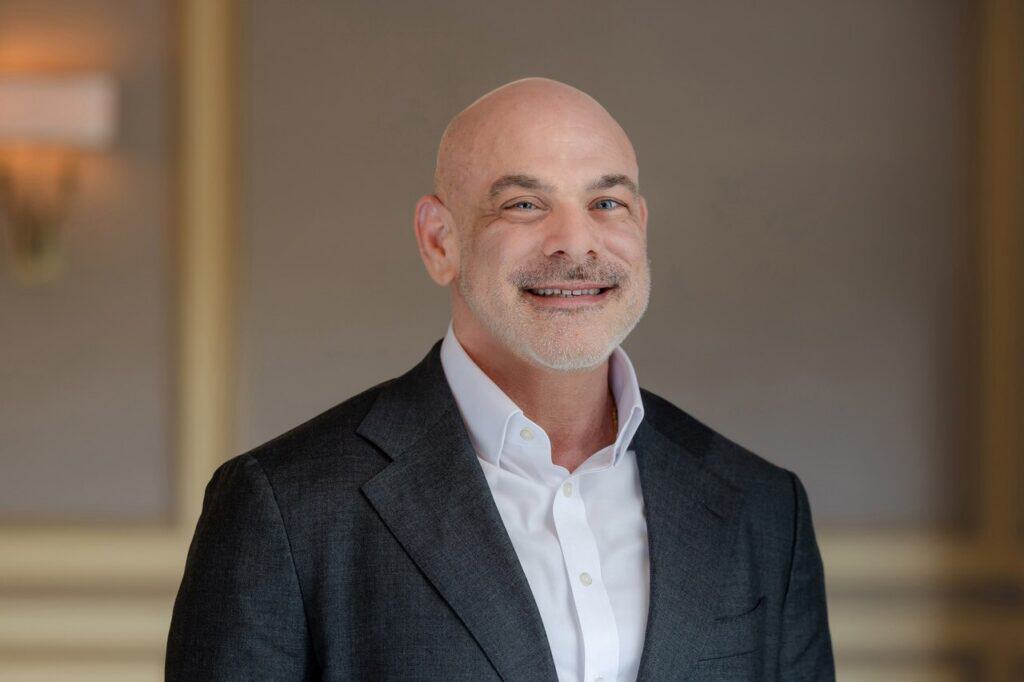 Headshot of a smiling bald man with a light beard, wearing a dark grey suit jacket and white shirt.