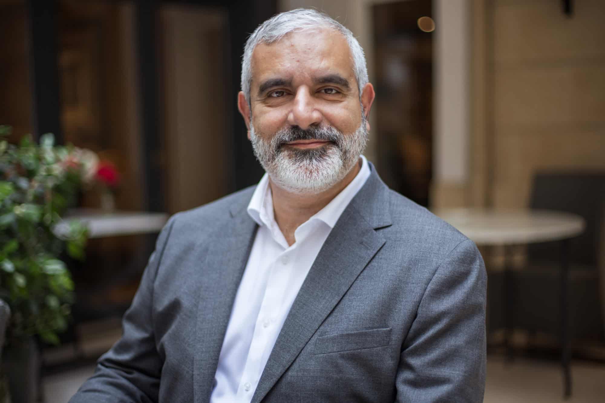 A man with gray hair and beard, wearing a gray suit and white shirt, smiles at the camera.