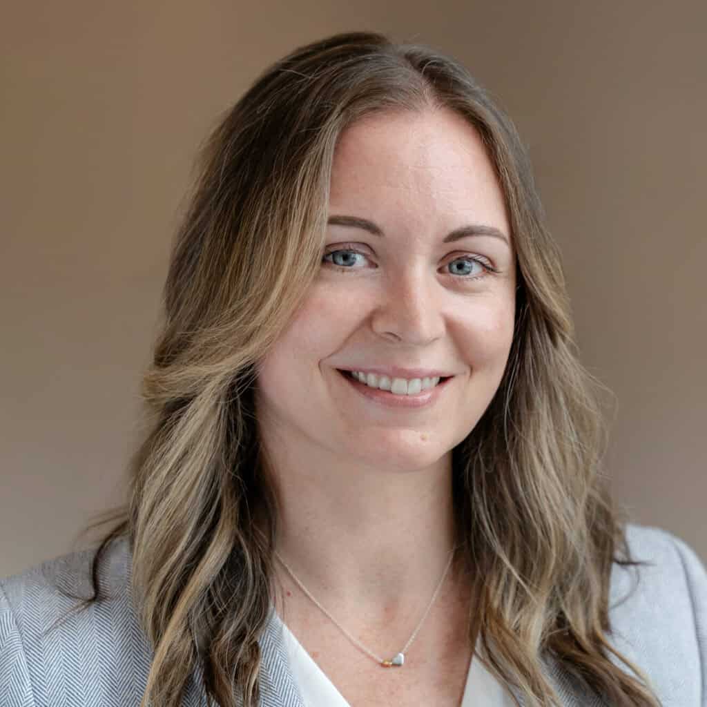 Headshot of a smiling woman with light brown hair, blue eyes, wearing a light blue herringbone blazer and a white V-neck top.