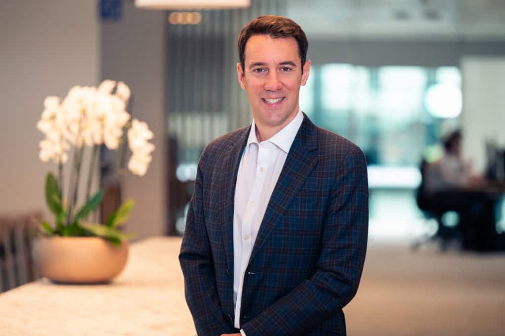 A smiling man in a blue plaid suit jacket and white shirt stands in an office setting with blurred white orchids in the background.