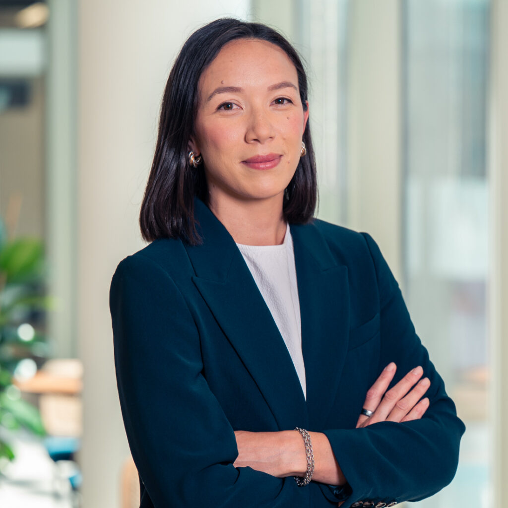 A woman with dark hair, wearing a teal blazer, stands with her arms crossed, smiling at the camera in a modern office.