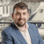 Headshot of a smiling man with light brown hair and beard, wearing a blue blazer and pink shirt, against a blurred urban background.