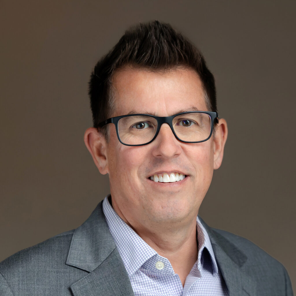 Headshot of a man with dark hair and black glasses, smiling in a gray suit jacket and checkered shirt.