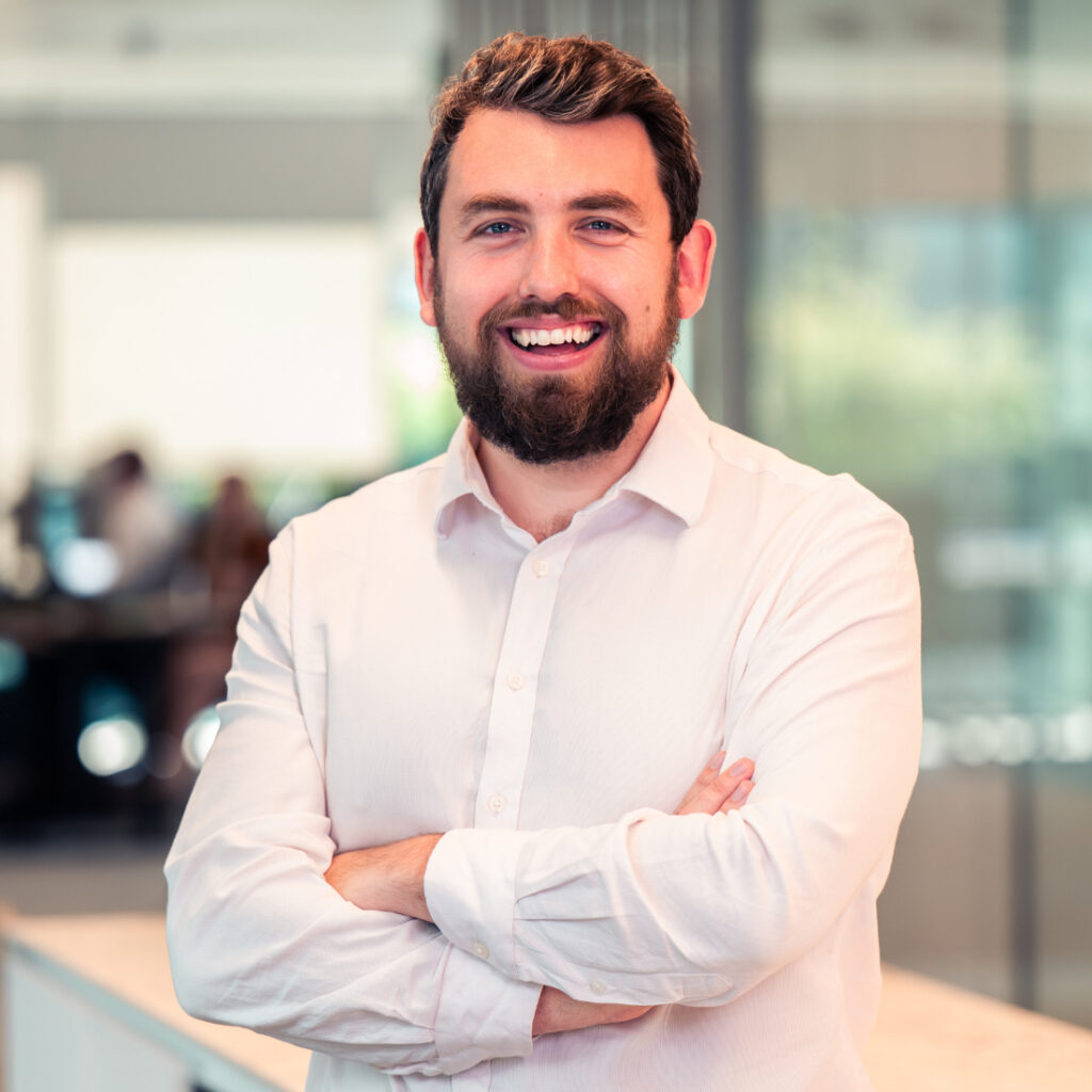 A man with a beard and a light shirt smiling with crossed arms in an office.