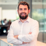 A man with a beard and a light shirt smiling with crossed arms in an office.