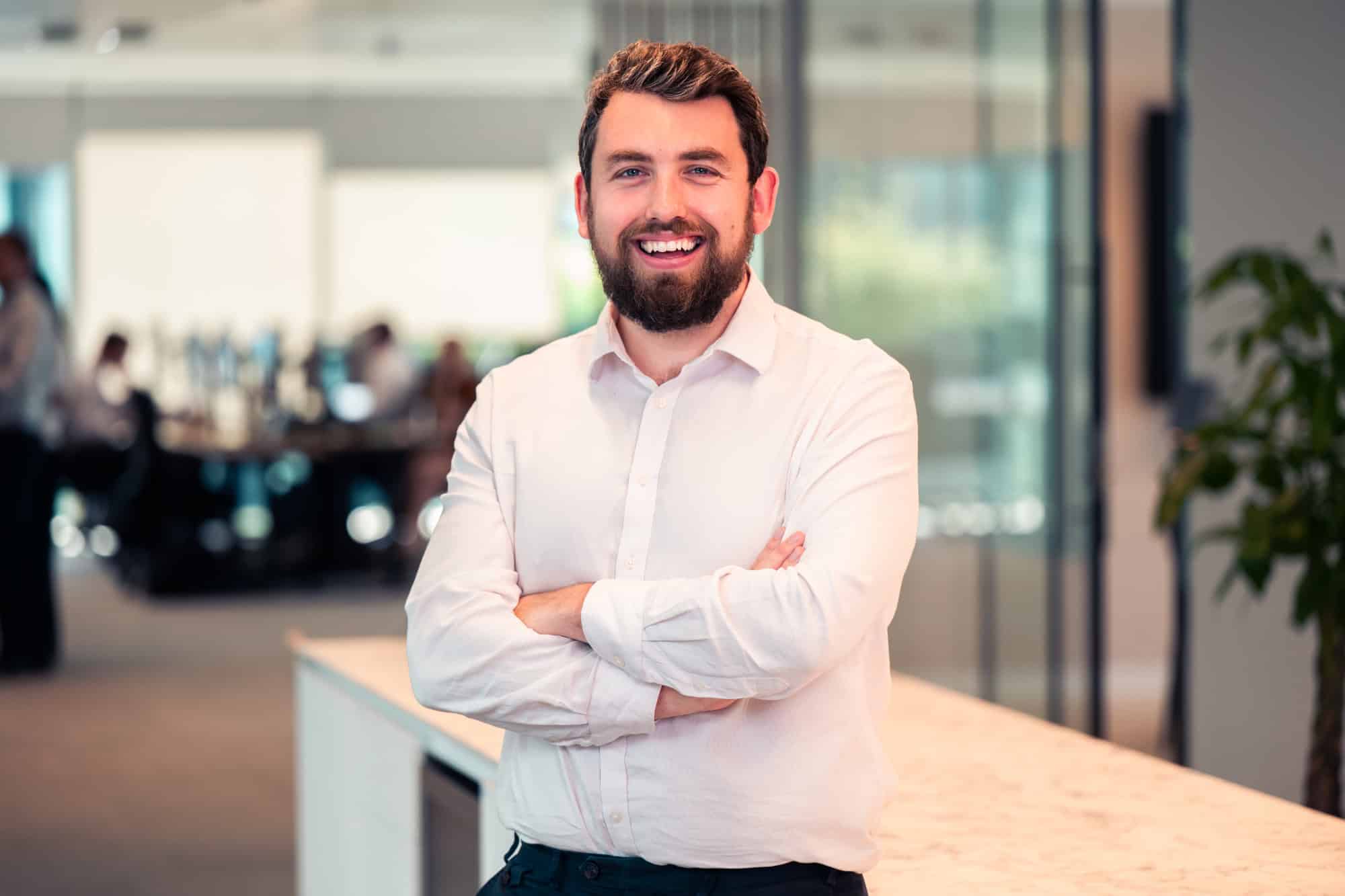 A man with a beard and a light shirt smiling with crossed arms in an office.