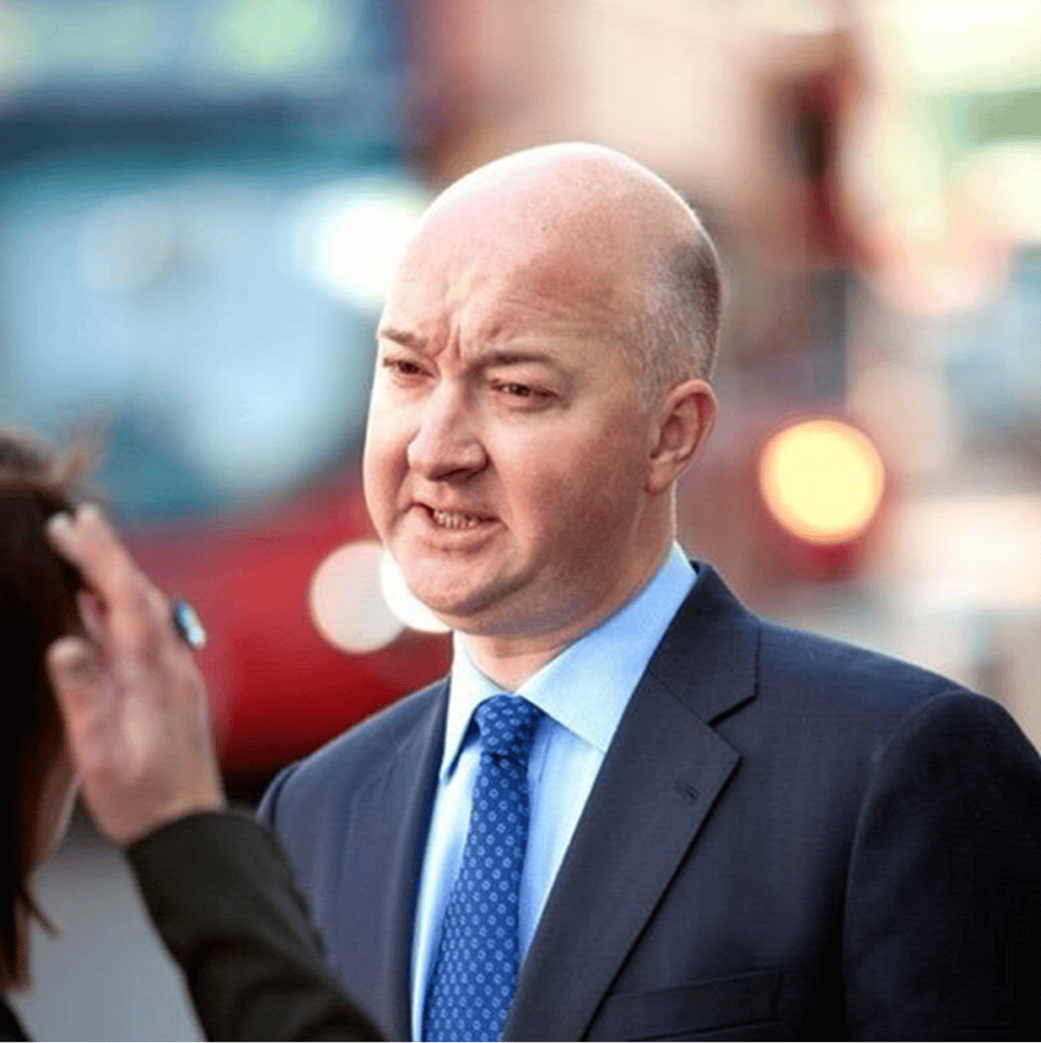 A bald man in a dark suit, light blue shirt, and blue tie, with a furrowed brow and open mouth, looking left. Part of another person's hand is visible on the left.