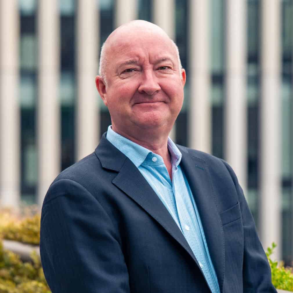 A bald man in a dark suit jacket and light blue shirt smiles at the camera, with green plants and a blurred modern building in the background.