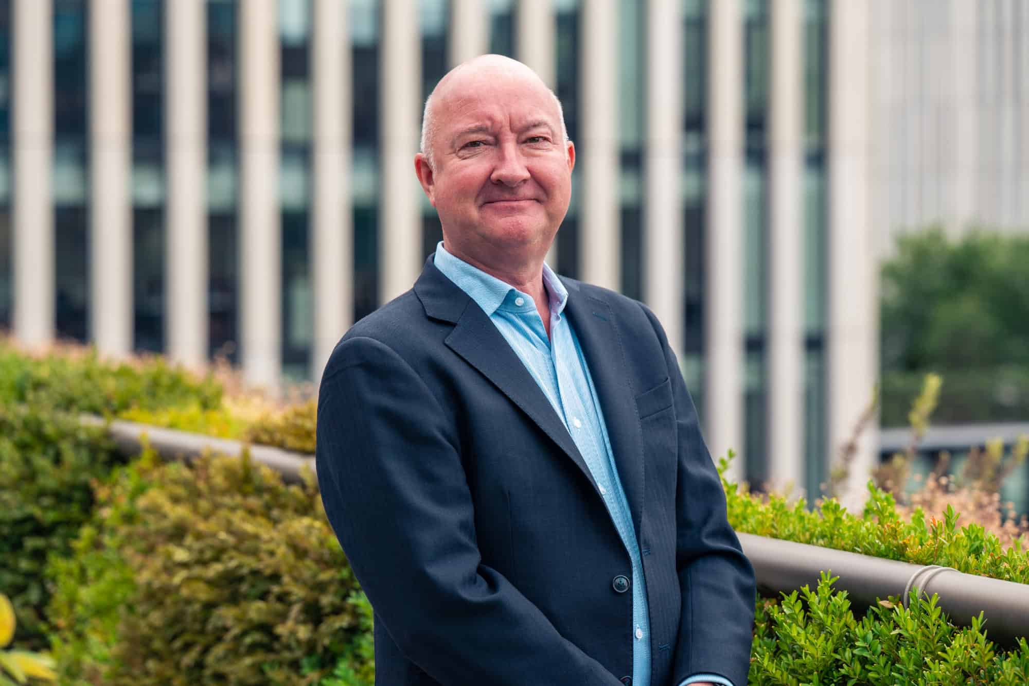 A bald man in a dark suit jacket and light blue shirt smiles at the camera, with green plants and a blurred modern building in the background.