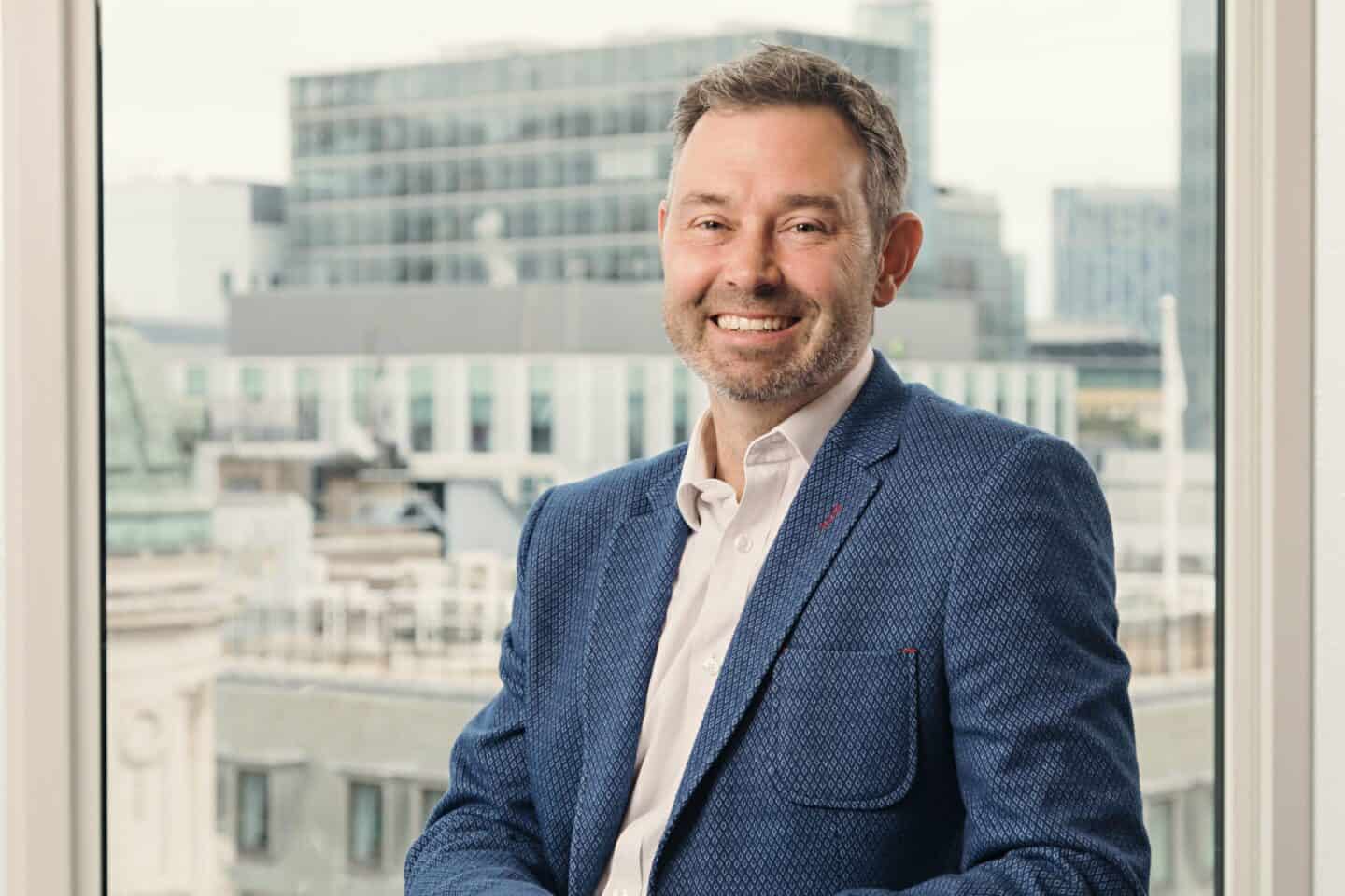 A man with a beard and short brown hair smiles, wearing a blue patterned blazer and a light shirt, with a blurred cityscape in the background.