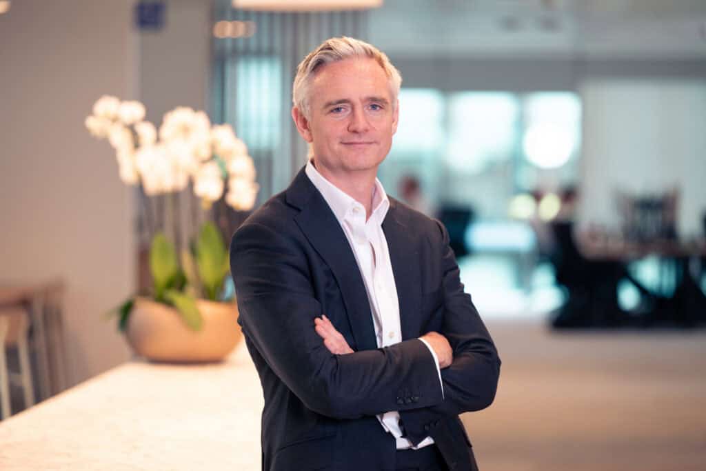 A man in a dark suit and white shirt with grey hair and blue eyes stands with arms crossed, smiling in a modern office.