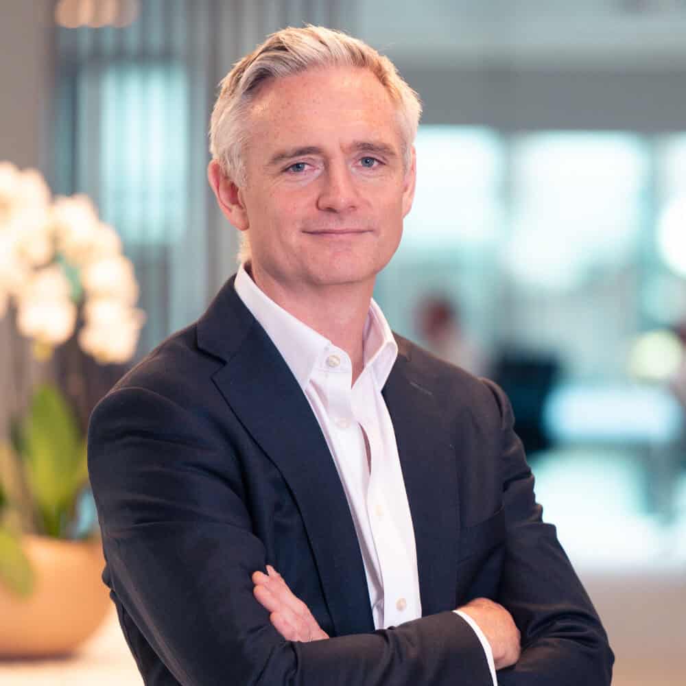 A man in a dark suit and white shirt with grey hair and blue eyes stands with arms crossed, smiling in a modern office.