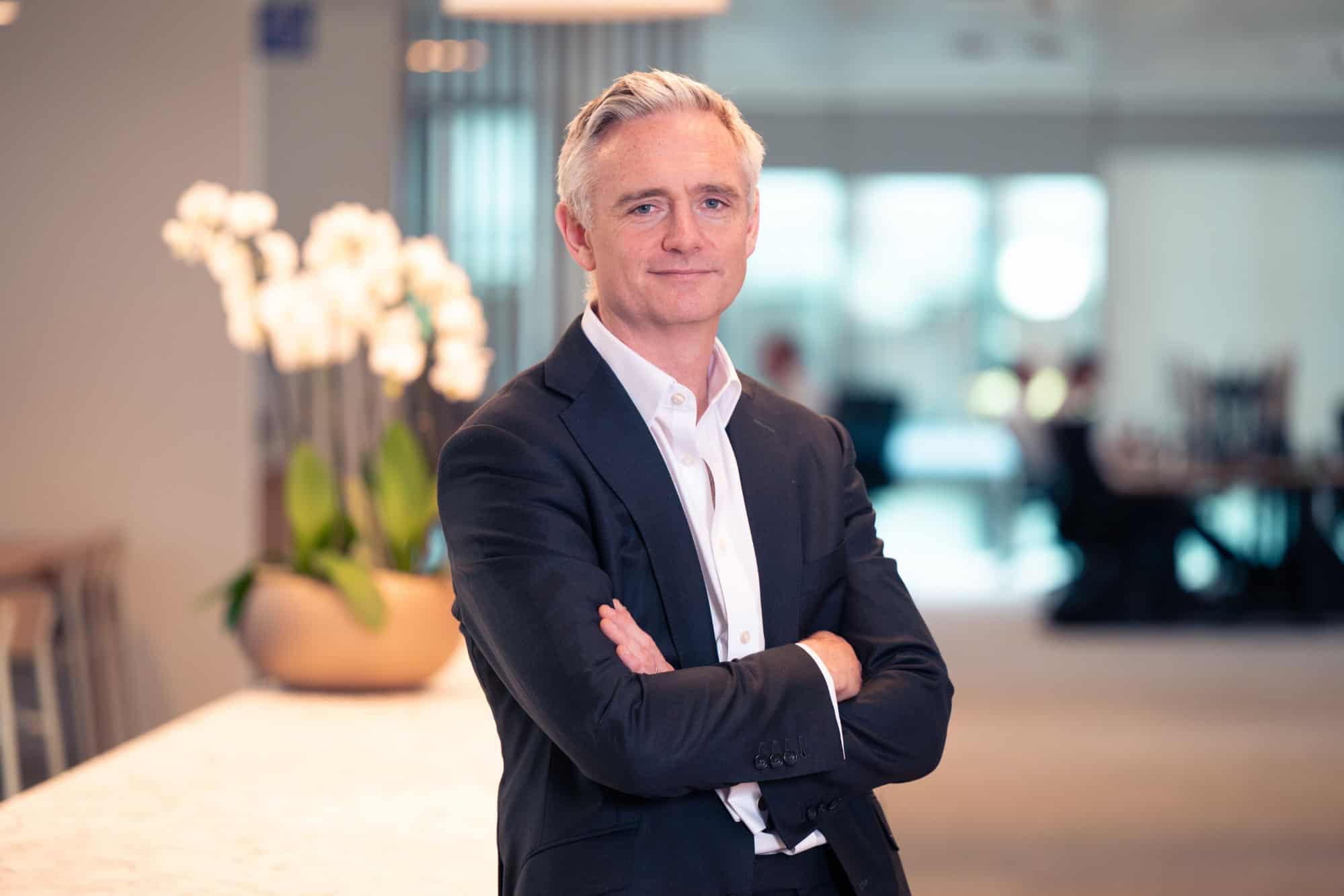 A man in a dark suit and white shirt with grey hair and blue eyes stands with arms crossed, smiling in a modern office.