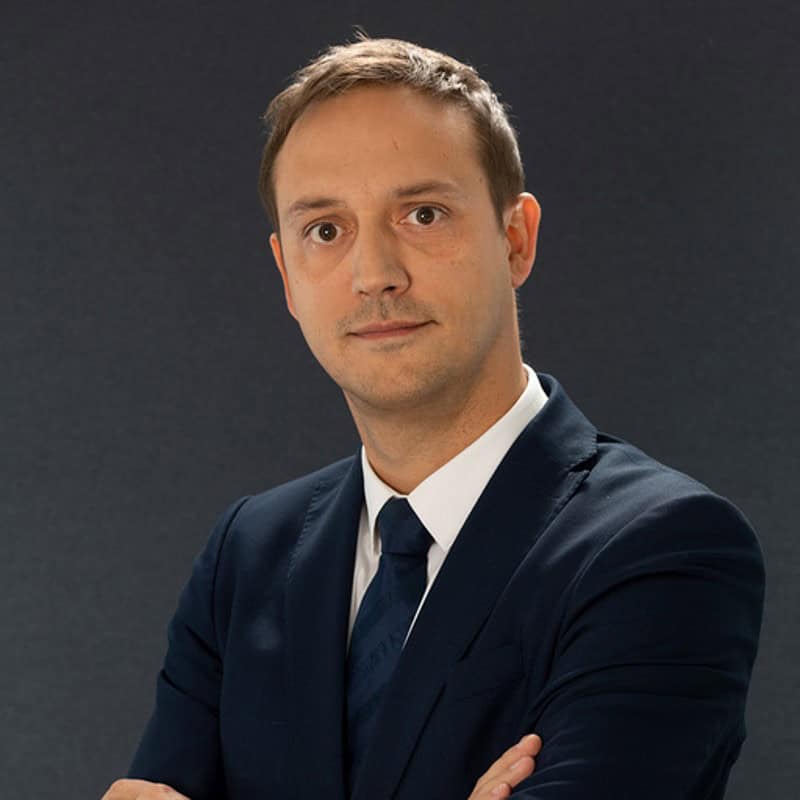 Headshot of a man in a navy blue suit and tie, with arms crossed, against a dark gray background.