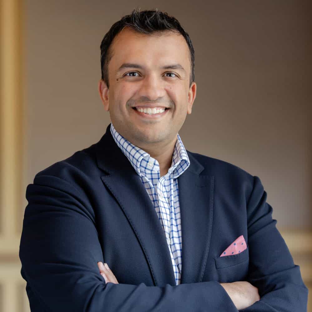 A smiling man in a navy blazer and checkered shirt with a pink pocket square, arms crossed, against a blurred warm-toned background with a wall sconce.