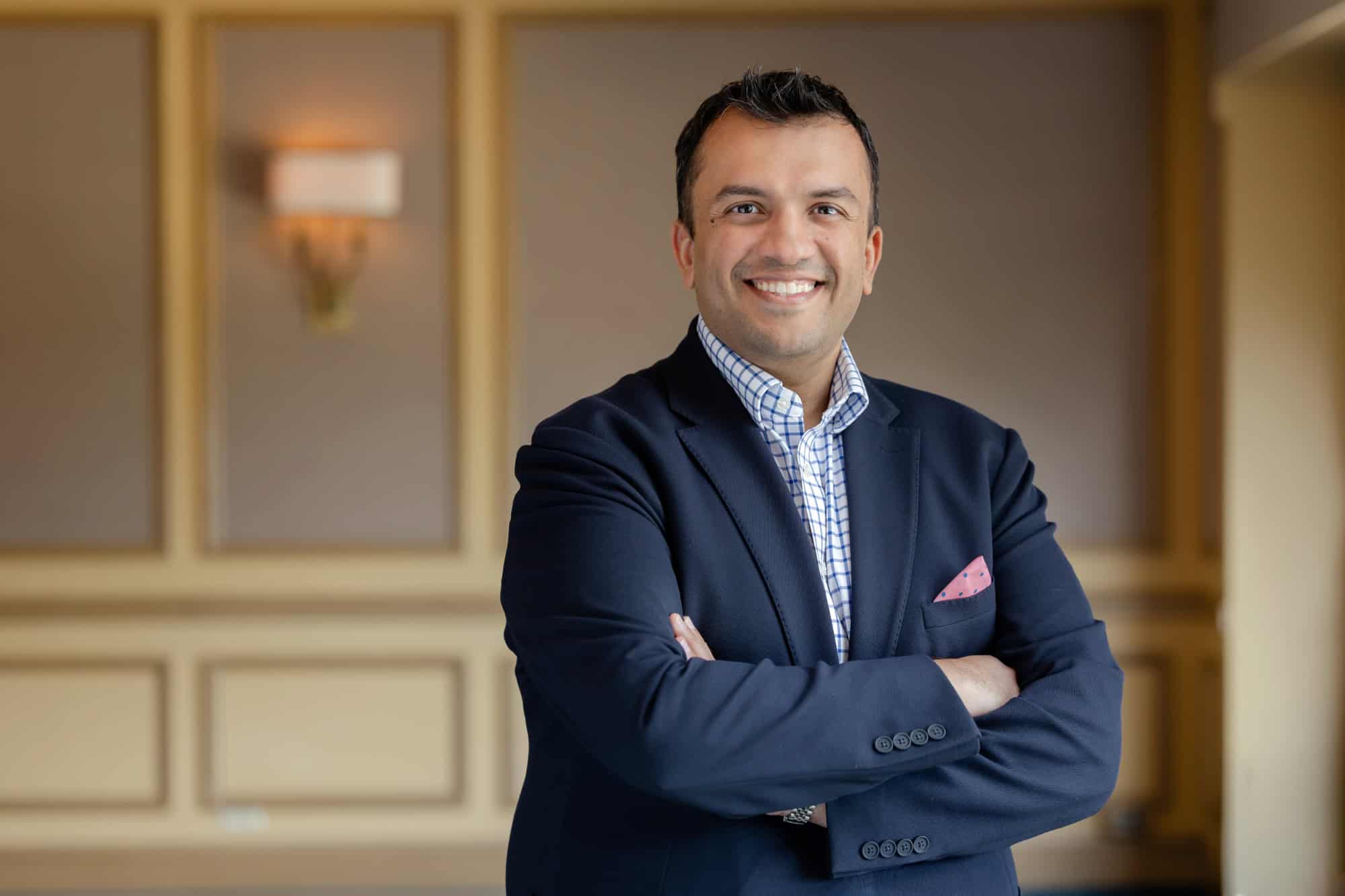 A smiling man in a navy blazer and checkered shirt with a pink pocket square, arms crossed, against a blurred warm-toned background with a wall sconce.