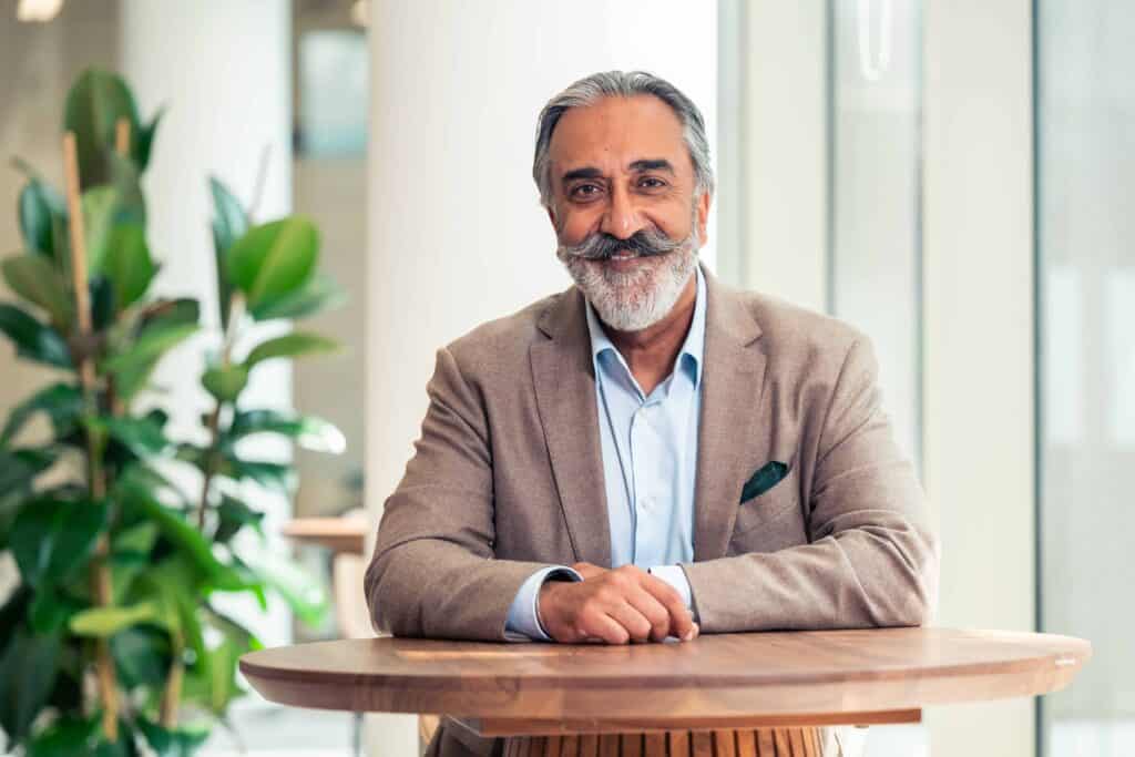 A smiling man with gray hair and a large mustache and beard, wearing a brown blazer and blue shirt, sits at a wooden table.
