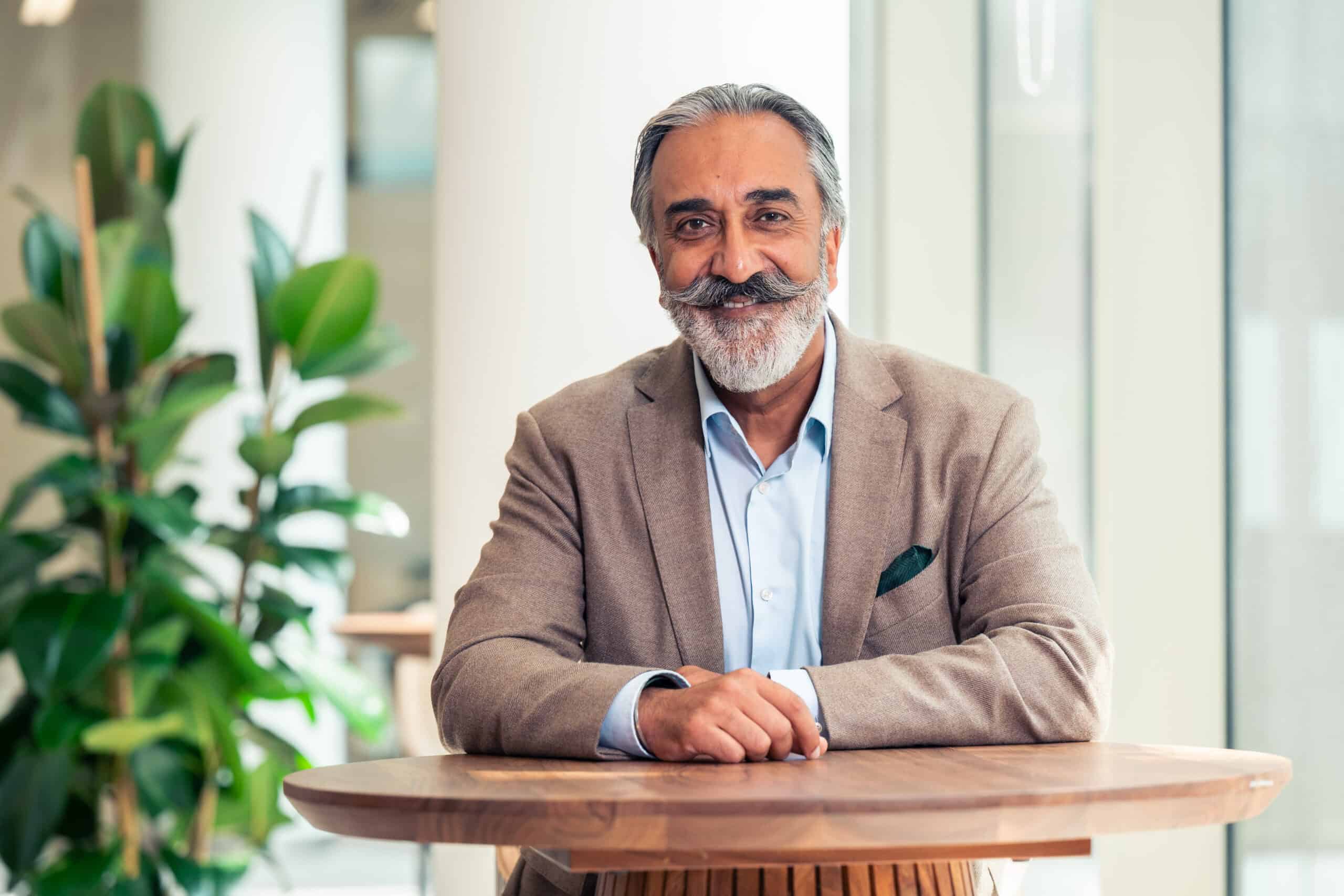 A smiling man with gray hair and a large mustache and beard, wearing a brown blazer and blue shirt, sits at a wooden table.