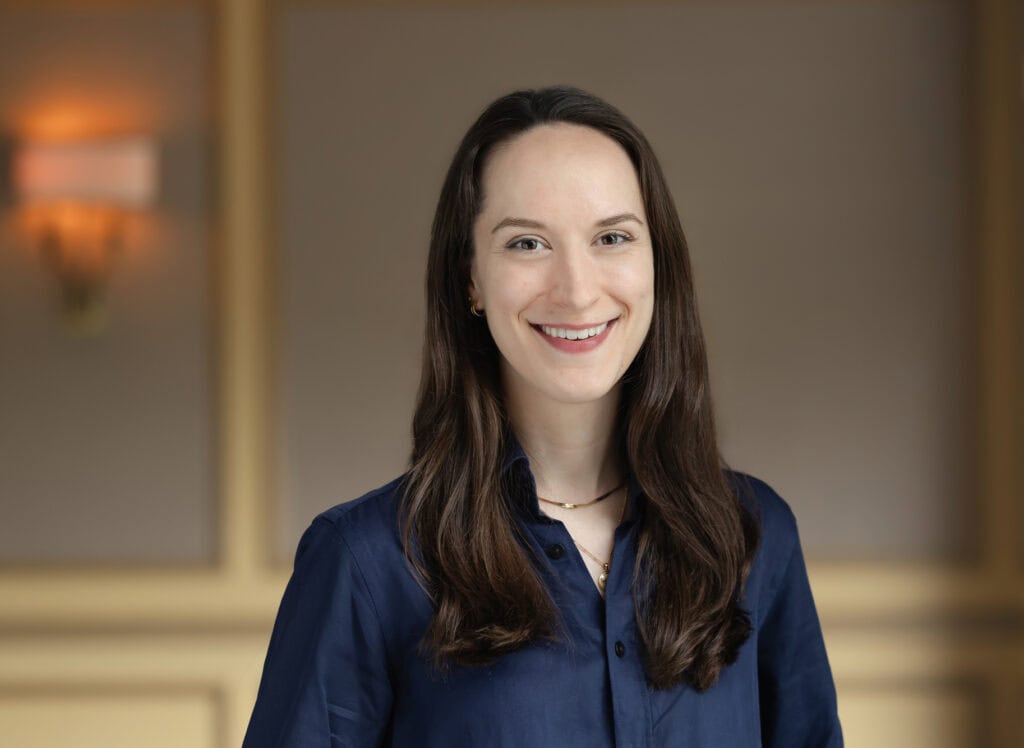 Headshot of a smiling young woman with long dark hair, wearing a blue shirt and gold necklaces, against a blurred warm background.