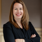 Headshot of a smiling woman with light brown hair, wearing a black blazer and a silver necklace, with arms crossed.