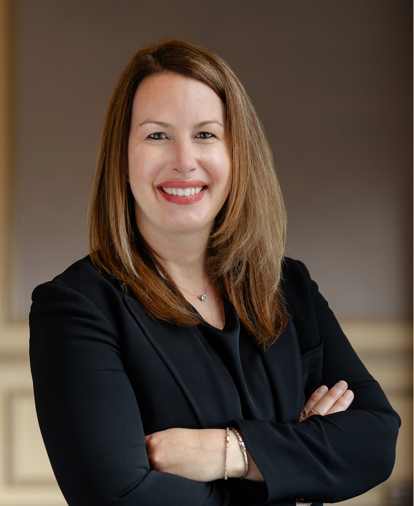 Headshot of a smiling woman with light brown hair, wearing a black blazer and a silver necklace, with arms crossed.