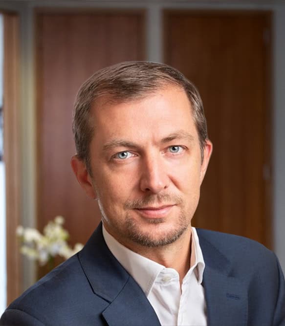Headshot of a man with short, graying hair, blue eyes, and light stubble, wearing a dark blue suit jacket and white shirt, looking at the camera.