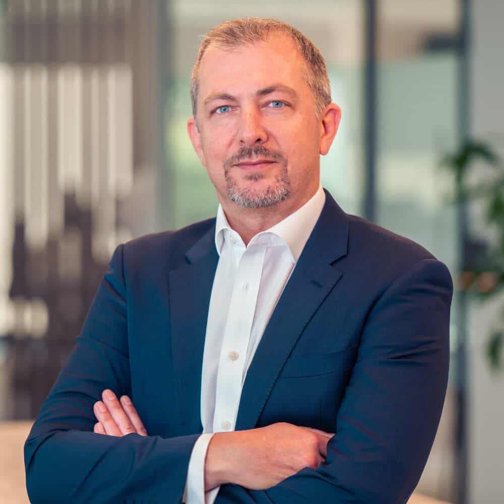 A man in a dark blue suit and white shirt, with graying hair and a beard, stands with arms crossed, looking at the camera in an office.