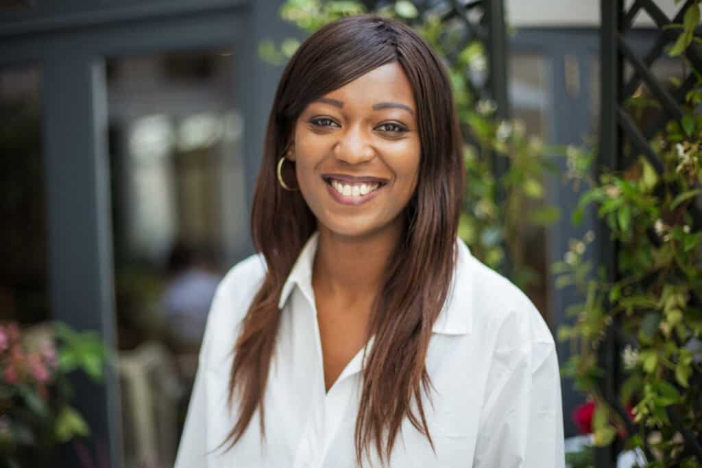 A smiling woman with dark skin and long dark hair wearing a white shirt and gold hoop earrings, looking at the camera.