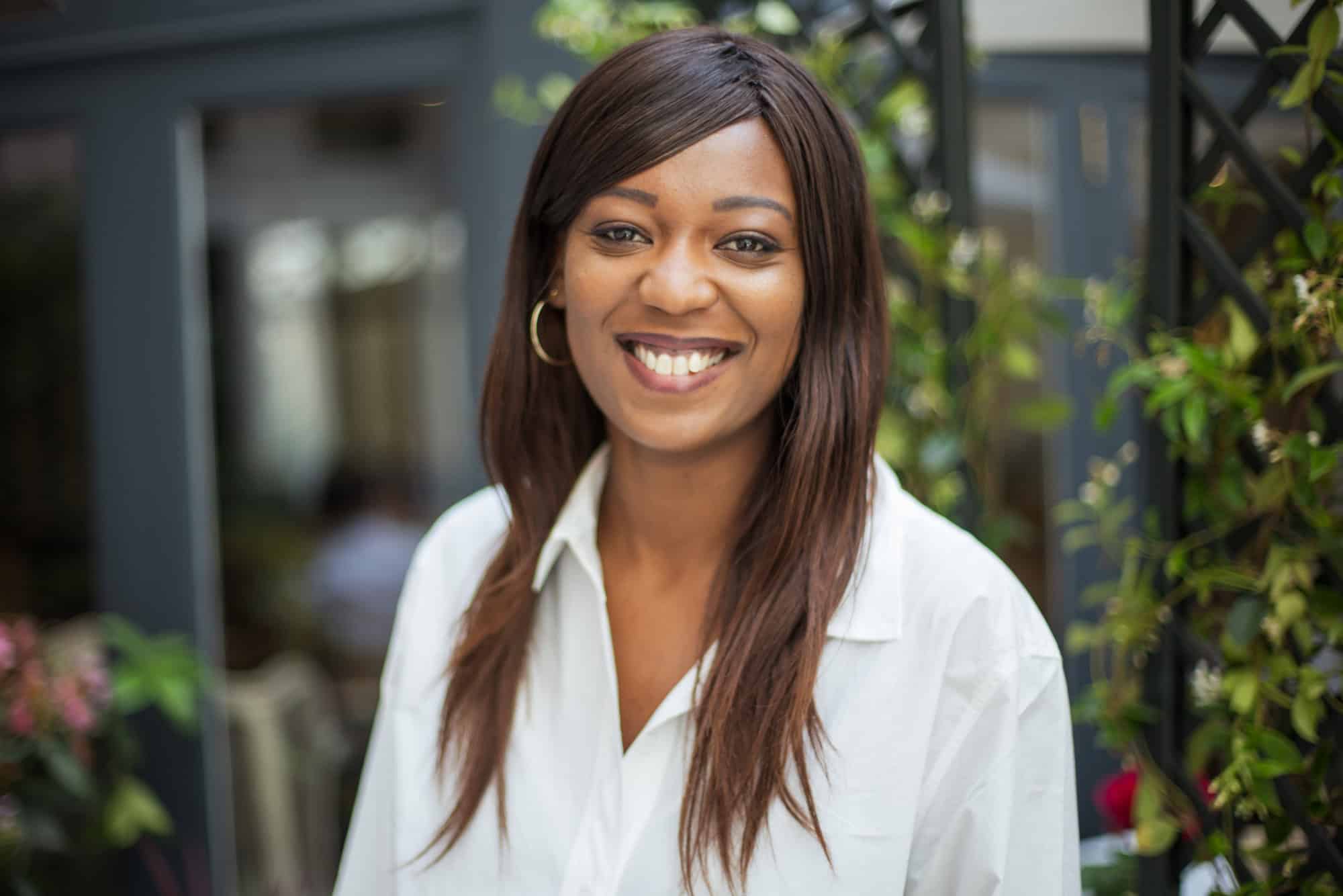 A smiling woman with dark skin and long dark hair wearing a white shirt and gold hoop earrings, looking at the camera.