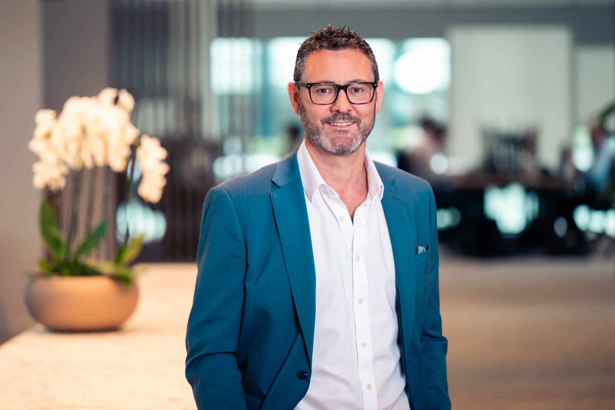 A man with dark hair, a grey beard, and glasses smiles, wearing a teal blazer and white shirt in a blurred indoor setting.