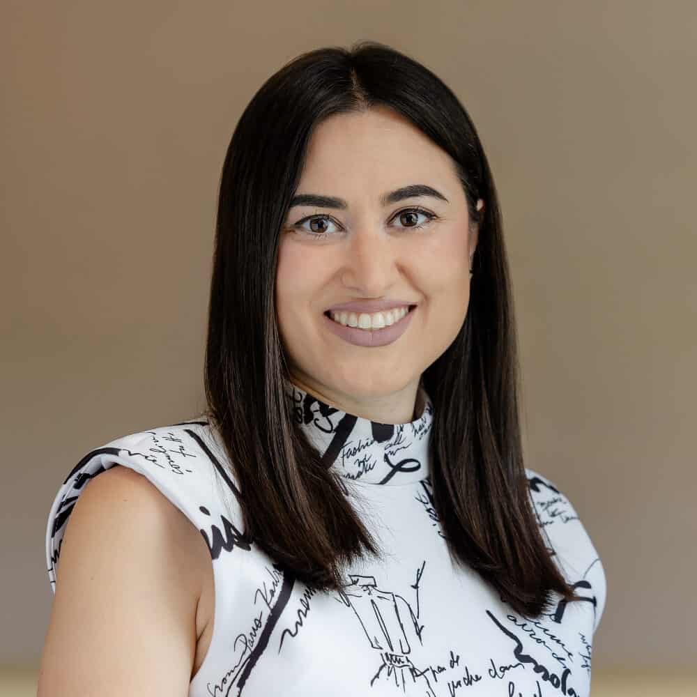 A smiling woman with dark hair and a white, script-print top looks at the camera.