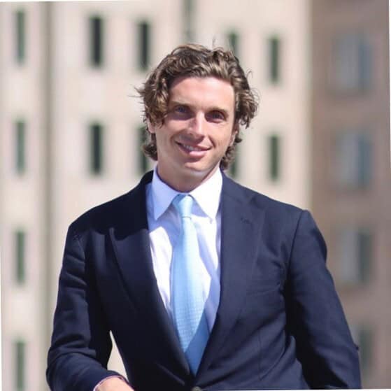 Headshot of a smiling man with curly brown hair, wearing a dark suit, white shirt, and light blue tie.