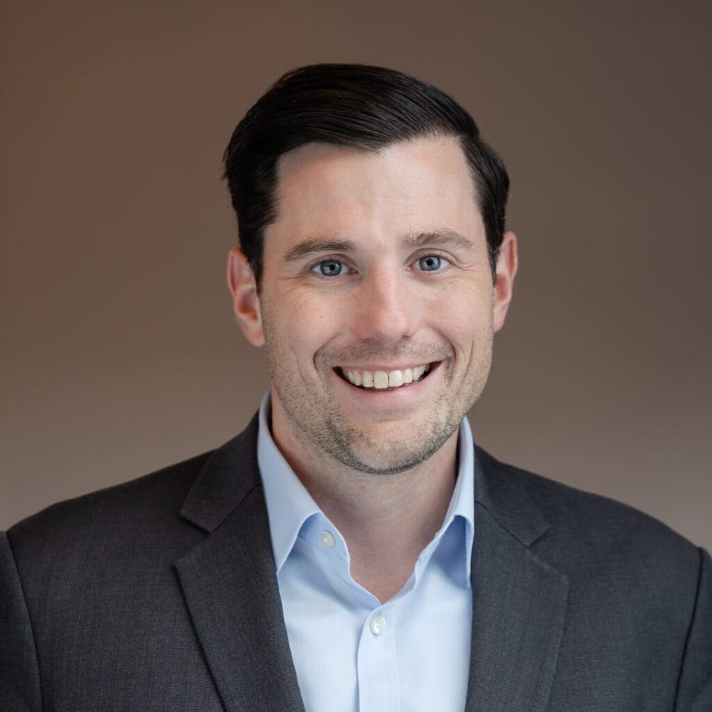 Headshot of a smiling man with dark hair, blue eyes, wearing a grey suit jacket and blue shirt.