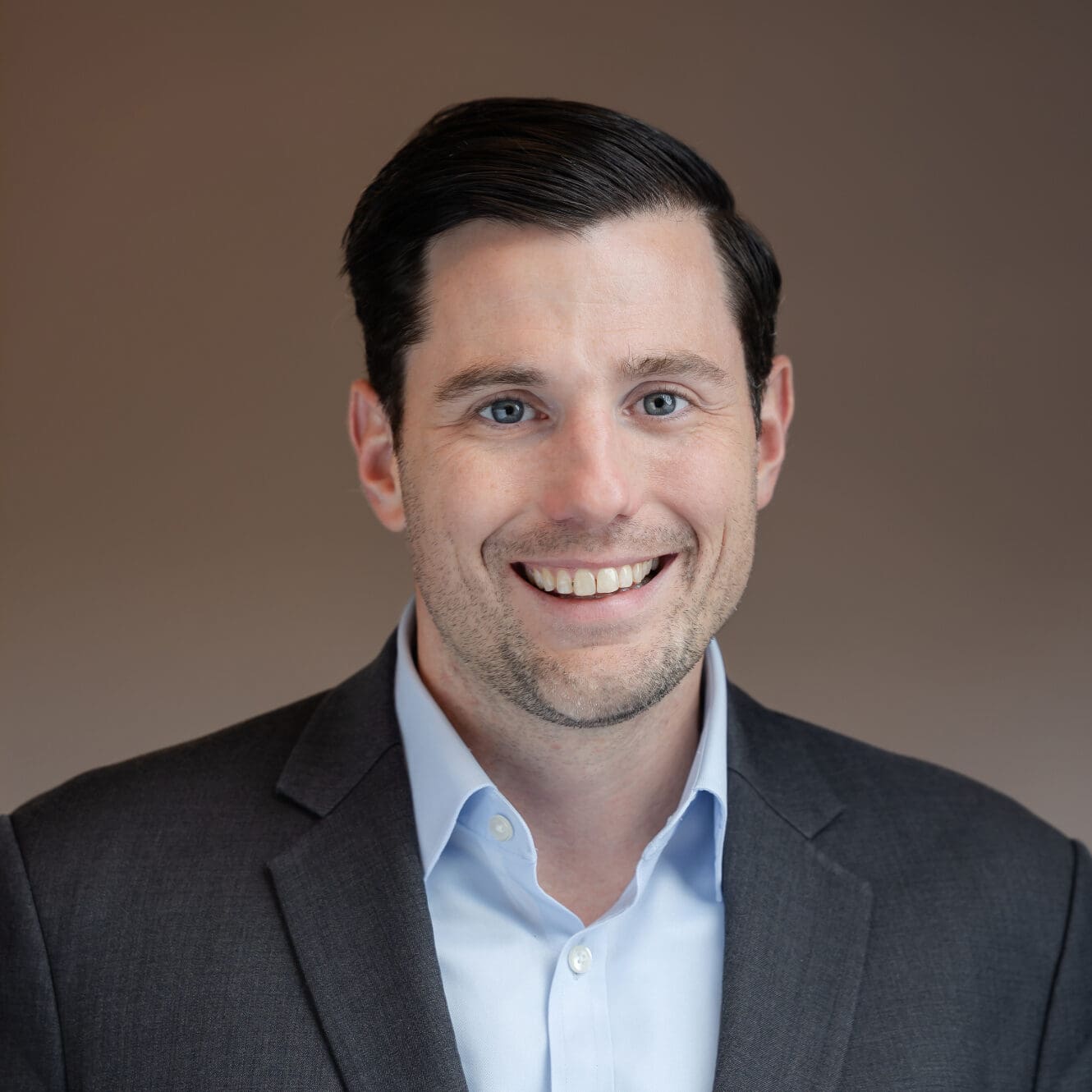 Headshot of a smiling man with dark hair, blue eyes, wearing a grey suit jacket and blue shirt.