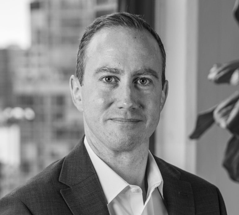 Black and white headshot of a man in a suit jacket and open-collared shirt, smiling slightly at the camera. Blurry city background.