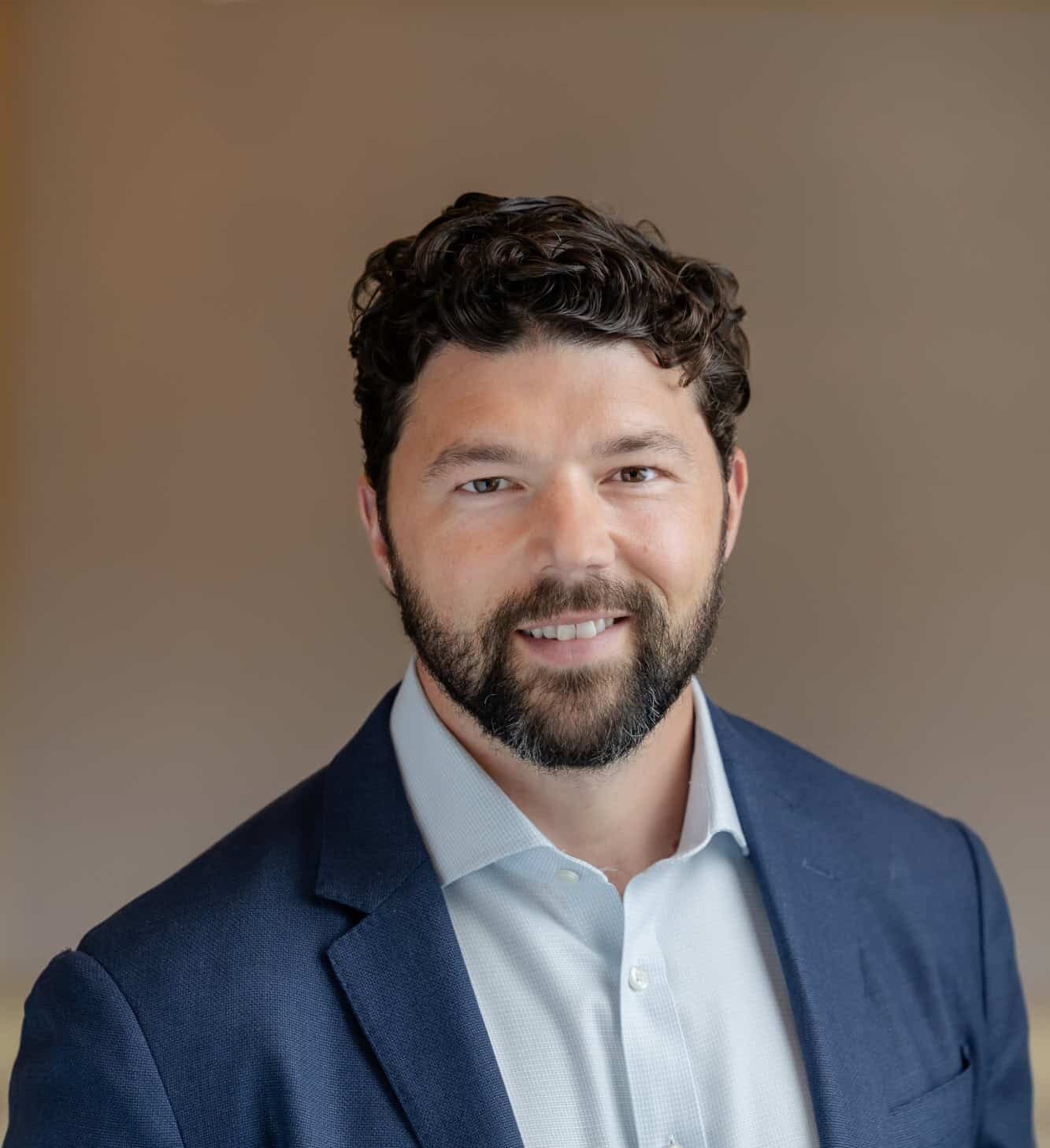 Headshot of a smiling man with dark curly hair and beard, wearing a blue blazer and light blue shirt.