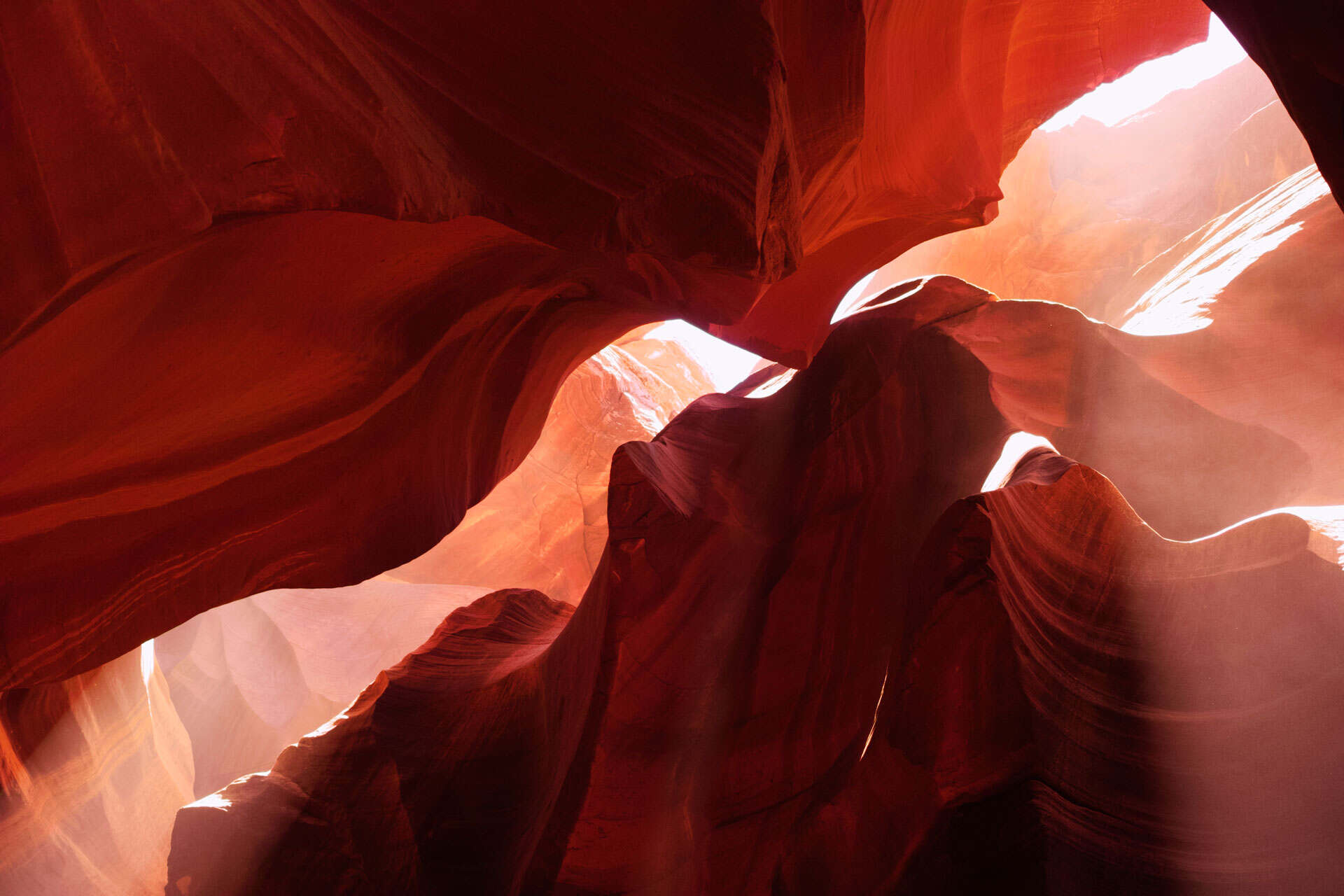 Interior view of a slot canyon with red and orange sandstone walls, illuminated by bright shafts of sunlight streaming from above.