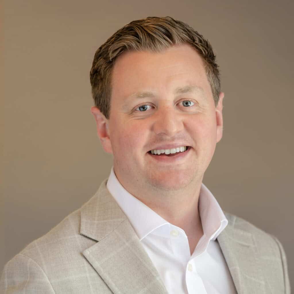 Headshot of a smiling man with light brown hair and blue eyes, wearing a light gray checked blazer and white shirt.