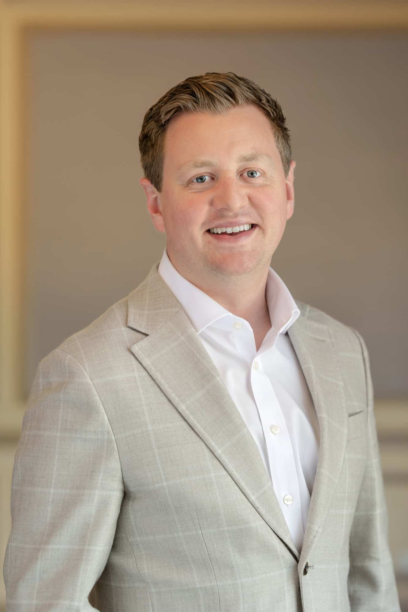 Headshot of a smiling man with light brown hair and blue eyes, wearing a light gray checked blazer and white shirt.