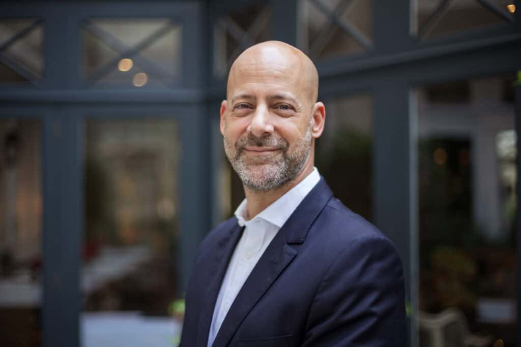 A bald man with a beard, wearing a dark suit and white shirt, smiles at the camera.