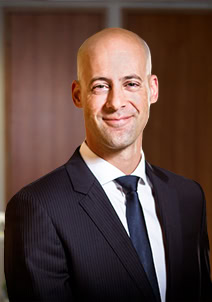 Headshot of a bald man in a dark pinstripe suit, white shirt, and dark blue tie, smiling.