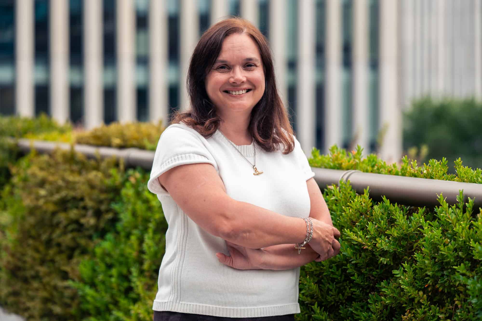 A smiling woman with dark hair and a white top stands with arms crossed in front of green bushes and a blurred building.