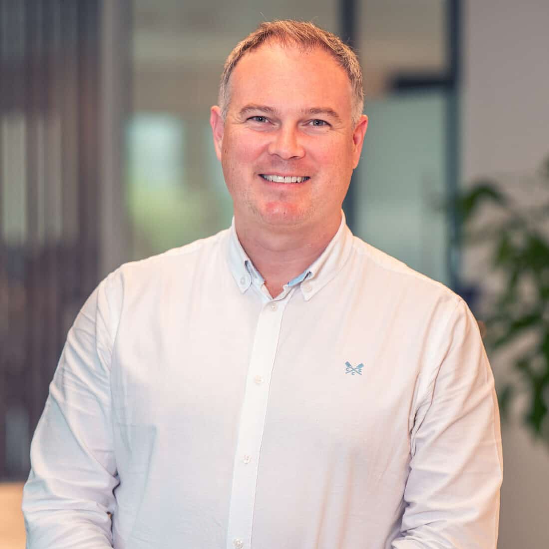 A man with short greying hair smiles at the camera, wearing a white button-down shirt with a logo, in an office setting.