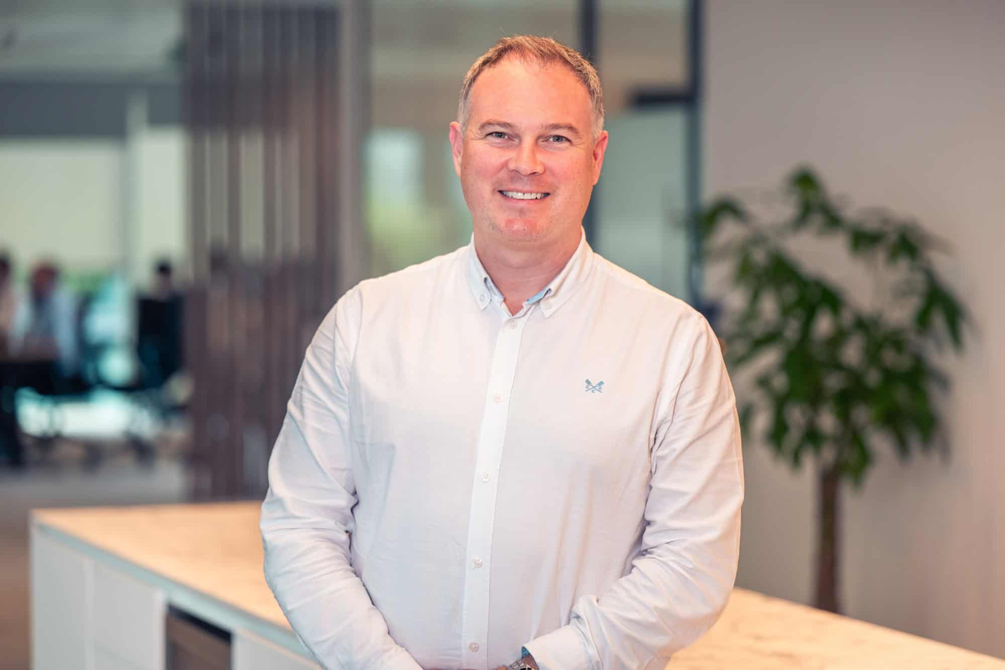 A man with short greying hair smiles at the camera, wearing a white button-down shirt with a logo, in an office setting.