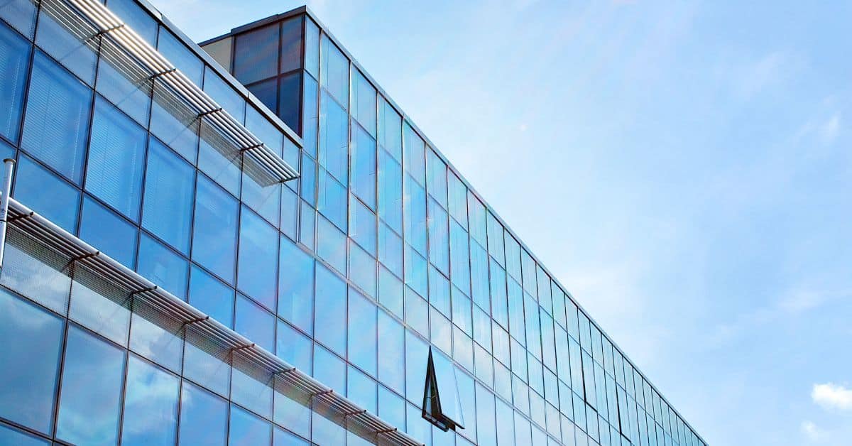 A modern building with a glass facade, featuring many rectangular windows, some with horizontal sunshades. One window is partially open. A clear blue sky is in the background.