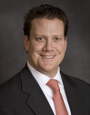 Headshot of a smiling man with dark hair, wearing a dark suit, white shirt, and red and white striped tie.