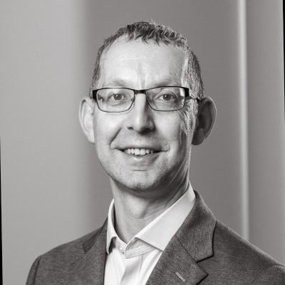 Black and white headshot of a smiling man with glasses, wearing a shirt and blazer.
