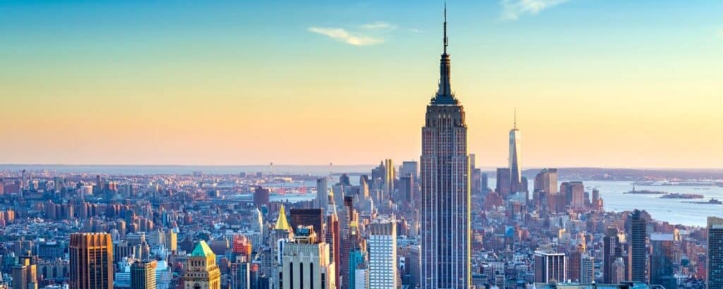 Panoramic view of the New York City skyline at sunset, with the Empire State Building and One World Trade Center prominent among many skyscrapers, and a body of water in the distance.